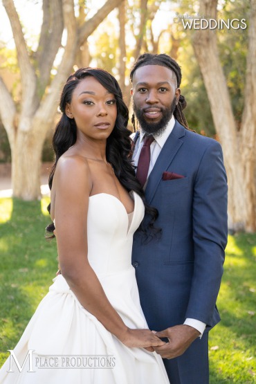 Stunning African American couple pose on their wedding dress. Bride with hair swept over one shoulder is wearing a white ballgown while the groom, holding her hand wears a cobalt blue tux and maroon tie.
