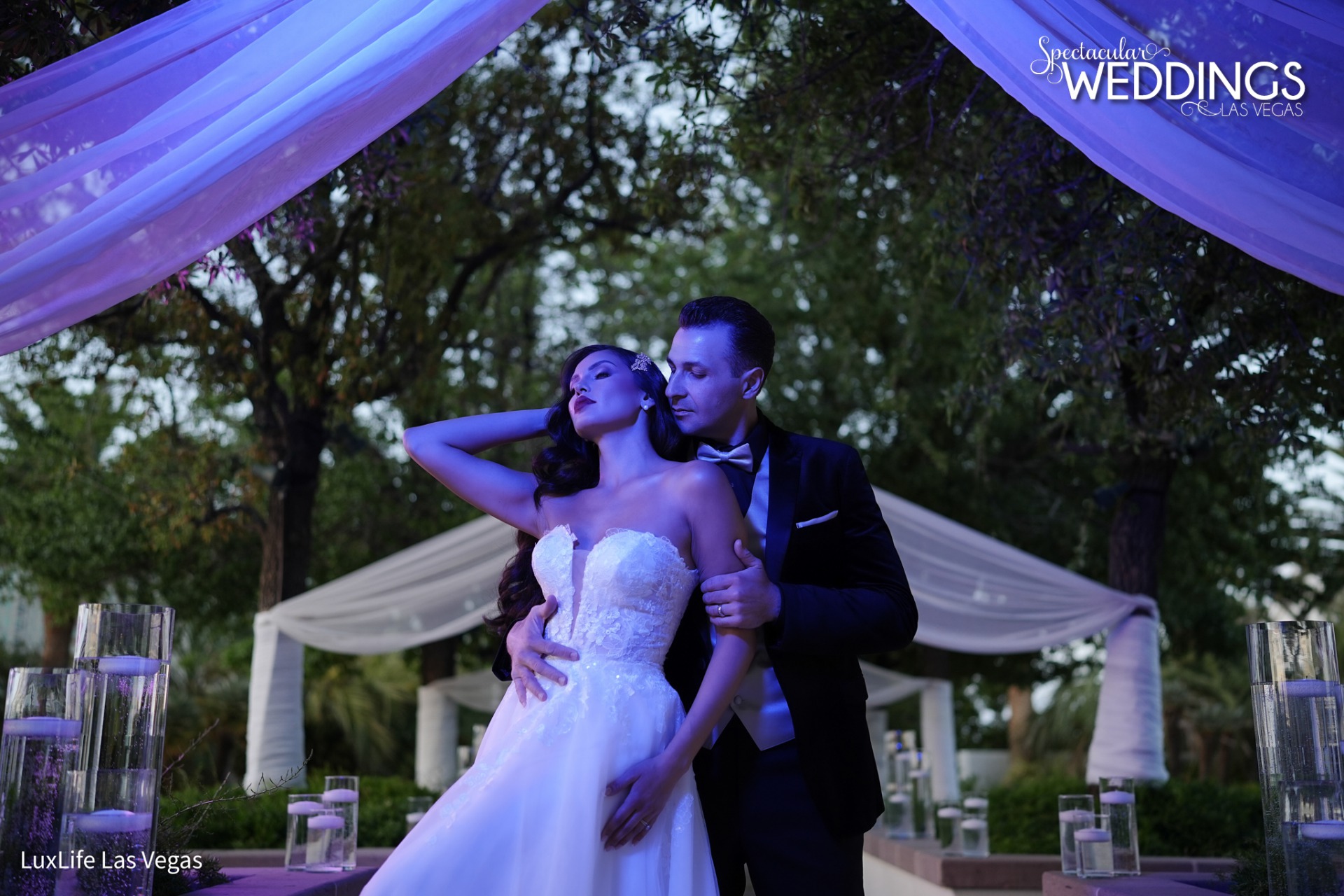 Romantic couple poses under white draped trees. Bride wears trending wedding gown while groom wears burgundy tuxedo.